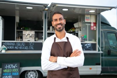 Mitarbeiter steht in Schürze vor seinem Food Truck und lächelt in die Kamera